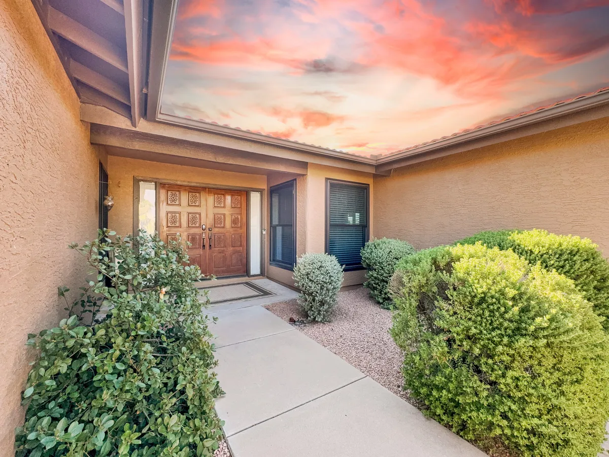 This exterior shot highlights an ornate wooden front door, beautifully landscaped shrubs, and a vibrant, colorful evening sky. The mood is inviting, perfect for welcoming gatherings and relaxed outdoor conversations as the day winds down.