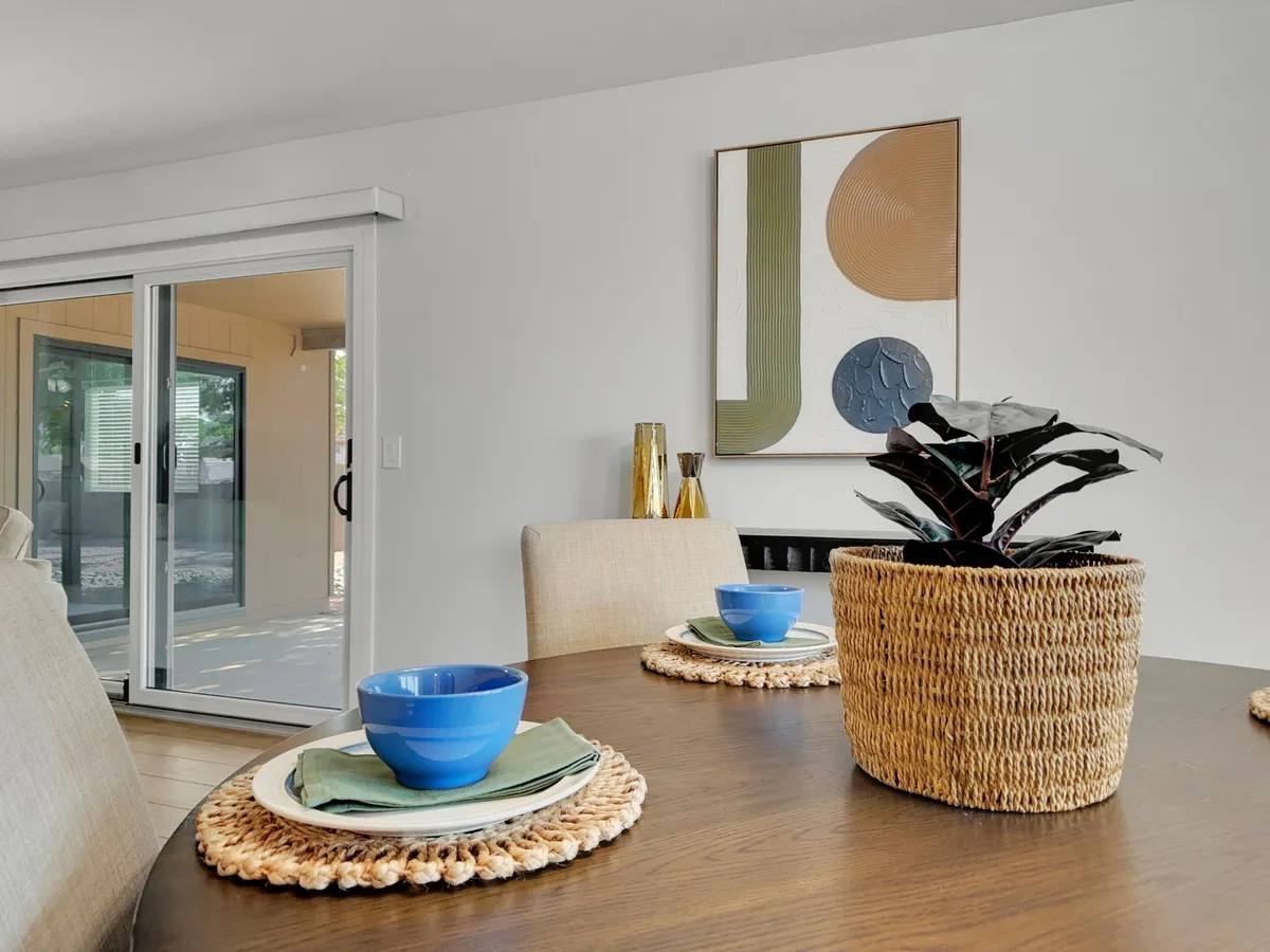 A cozy dining area with a dining table set for a meal, complemented by wall art, a plant in a basket, and blue bowls. The ambiance is warm and welcoming, ideal for family breakfasts or casual dining.