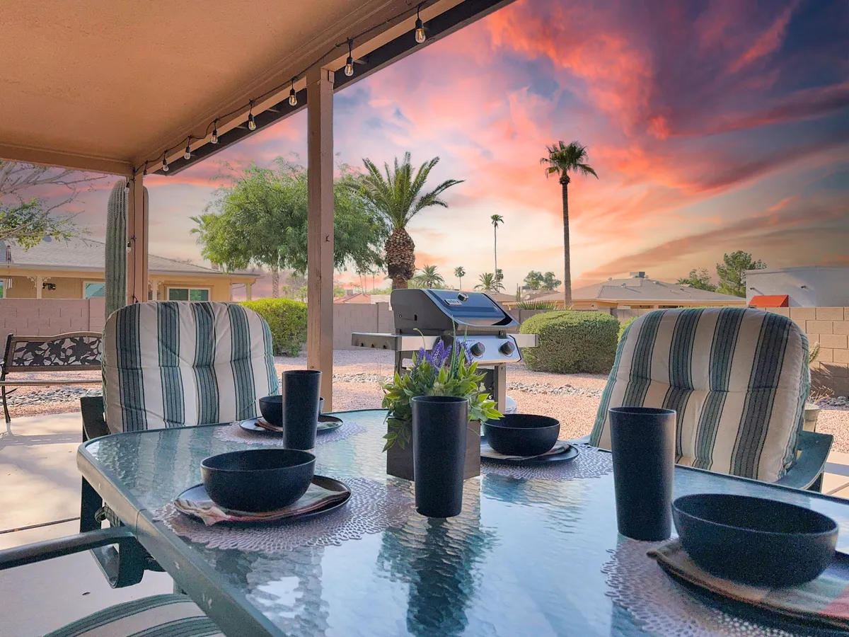 An inviting outdoor dining setup is pictured with a table set for a meal, a grill in the background, tall palm trees adding a resort-like vibe, and a colorful Arizona sunset painting the sky. The mood is cheerful and relaxed, hinting at memorable sunset dinners and evenings under the stars.
