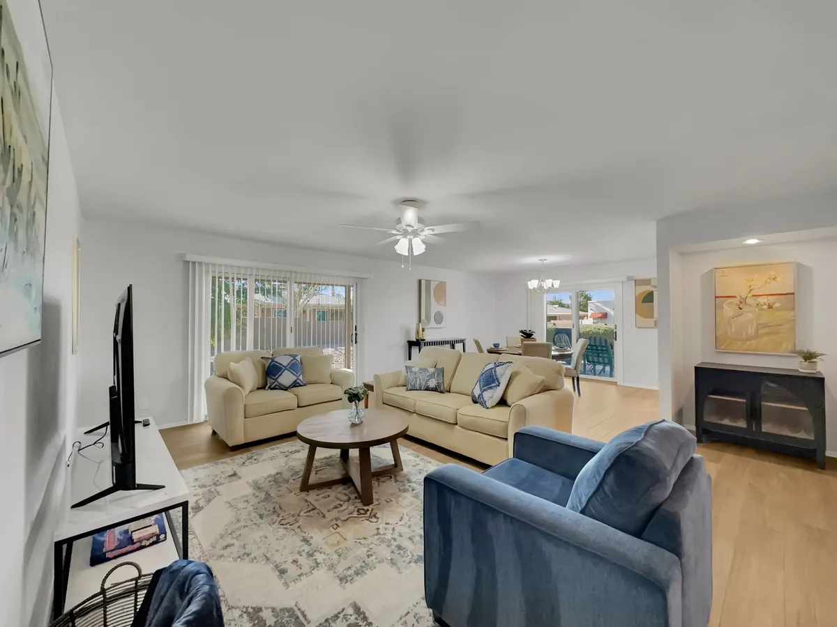 A spacious and inviting living room featuring a plush sofa set, a modern coffee table, tasteful artwork adorning the wall, a large television, and a window with blinds allowing soft natural light to filter in. The arrangement encourages group gatherings and comfortable relaxation.